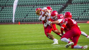 Several american football players on the field. One has the ball and is being tackled by two opponents.