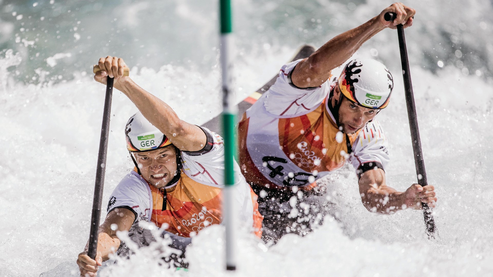 Two canoe athletes in the river, paddling between the poles, lots of water around them.