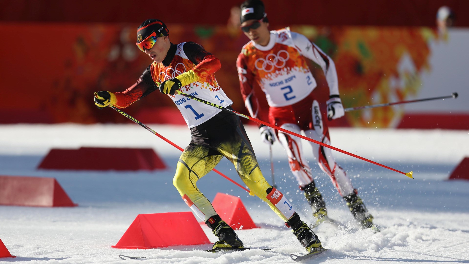 Two skiers in an Olympic competition about to make a right turn, both standing up, poles to the side.