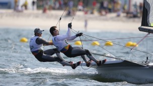 Two athletes in a sailingcompetito, both athletes standing and hanging on the side of the boar, holding on to the ropes.